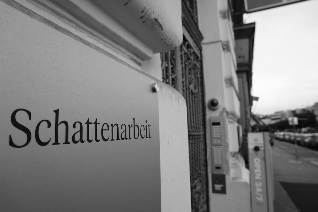 Foto eines Messingsschilds an einer Hauswand. Man erkennt das Wort "Schattenarbeit".